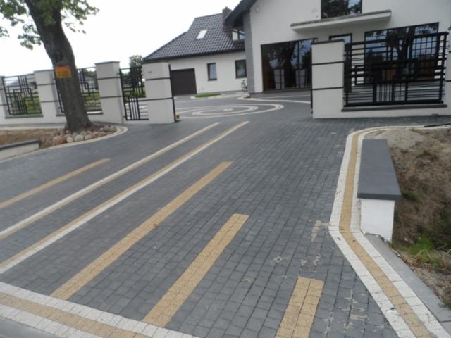 Szara kostka brukowa z żółtymi pasami prowadząca do domu z bramą i ogrodzeniem. Okrągły wzór z kostki przed budynkiem. Widok na posesję z zewnątrz.