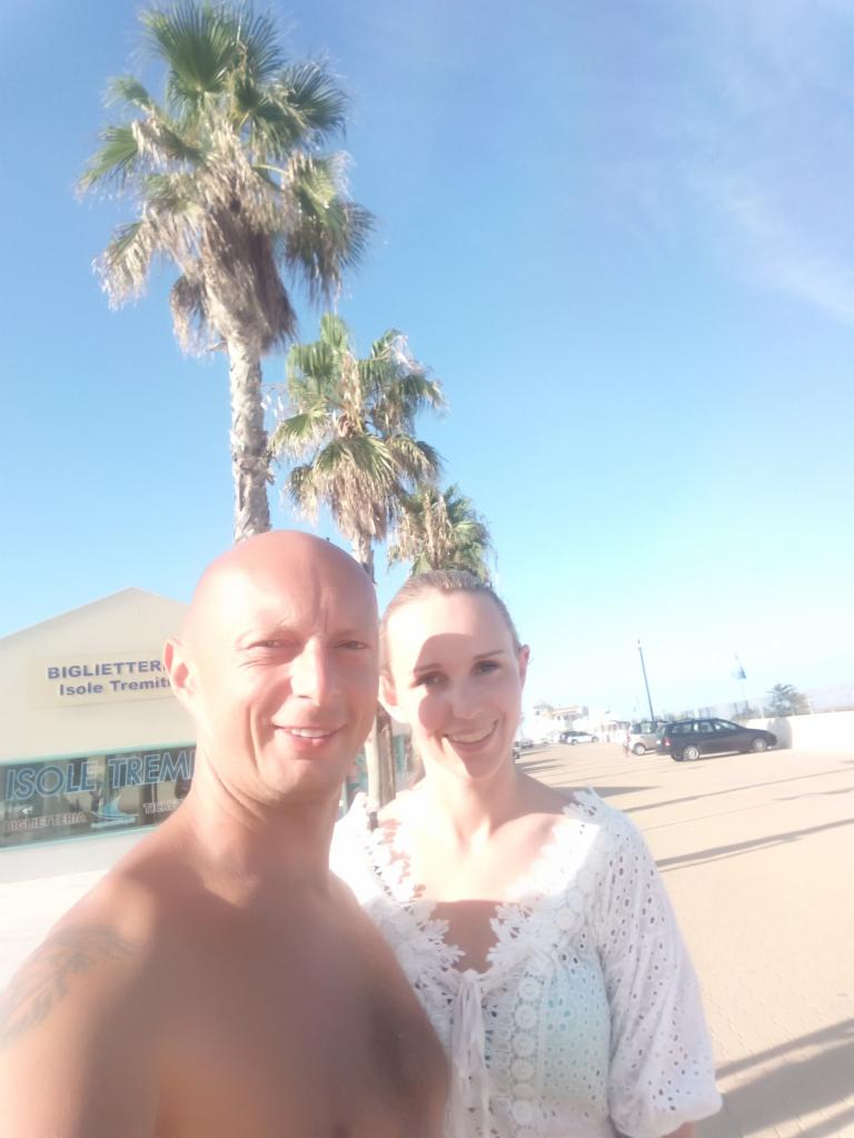 Uśmiechnięta para na tle palm i błękitnego nieba w słoneczny dzień. Widoczny budynek z napisem 'Isole Tremiti' i zaparkowane samochody w tle.