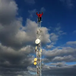Mężczyzna w czerwonej kurtce na szczycie wieży telekomunikacyjnej z antenami na tle pochmurnego nieba. Kąt ujęcia od dołu, podkreślający wysokość konstrukcji.