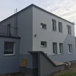 Dwukondygnacyjny budynek po malowaniu elewacji na dwa odcienie szarości; widoczna drabina na dachu, skrzynka pocztowa i zejście do piwnicy.