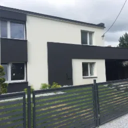 Dwukondygnacyjny dom z nowoczesną elewacją w kolorach bieli i antracytu, widoczny zza metalowego ogrodzenia panelowego.