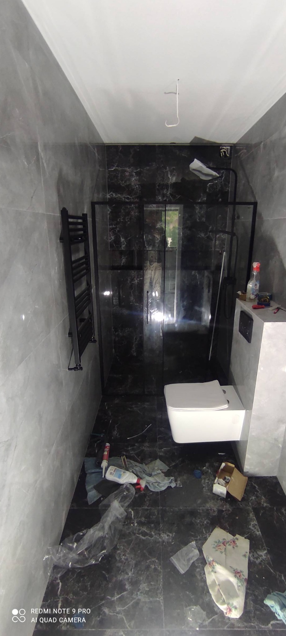 Wnętrze łazienki w trakcie remontu z czarno-szarymi marmurkowymi płytkami, widoczna kabina prysznicowa z czarnymi ramami, wisząca toaleta, grzejnik drabinkowy i porozrzucane narzędzia...