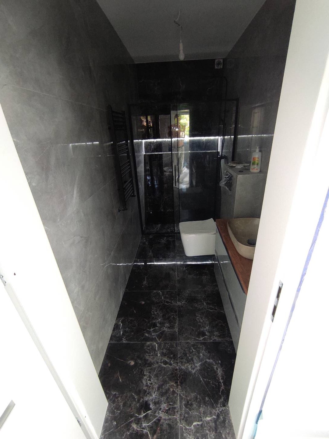 Wykończona łazienka z ciemnymi marmurkowymi płytkami na podłodze i ścianach, kabina prysznicowa z czarnymi profilami, widoczny fragment umywalki i toaleta, surowa żarówka zwisa z sufitu.