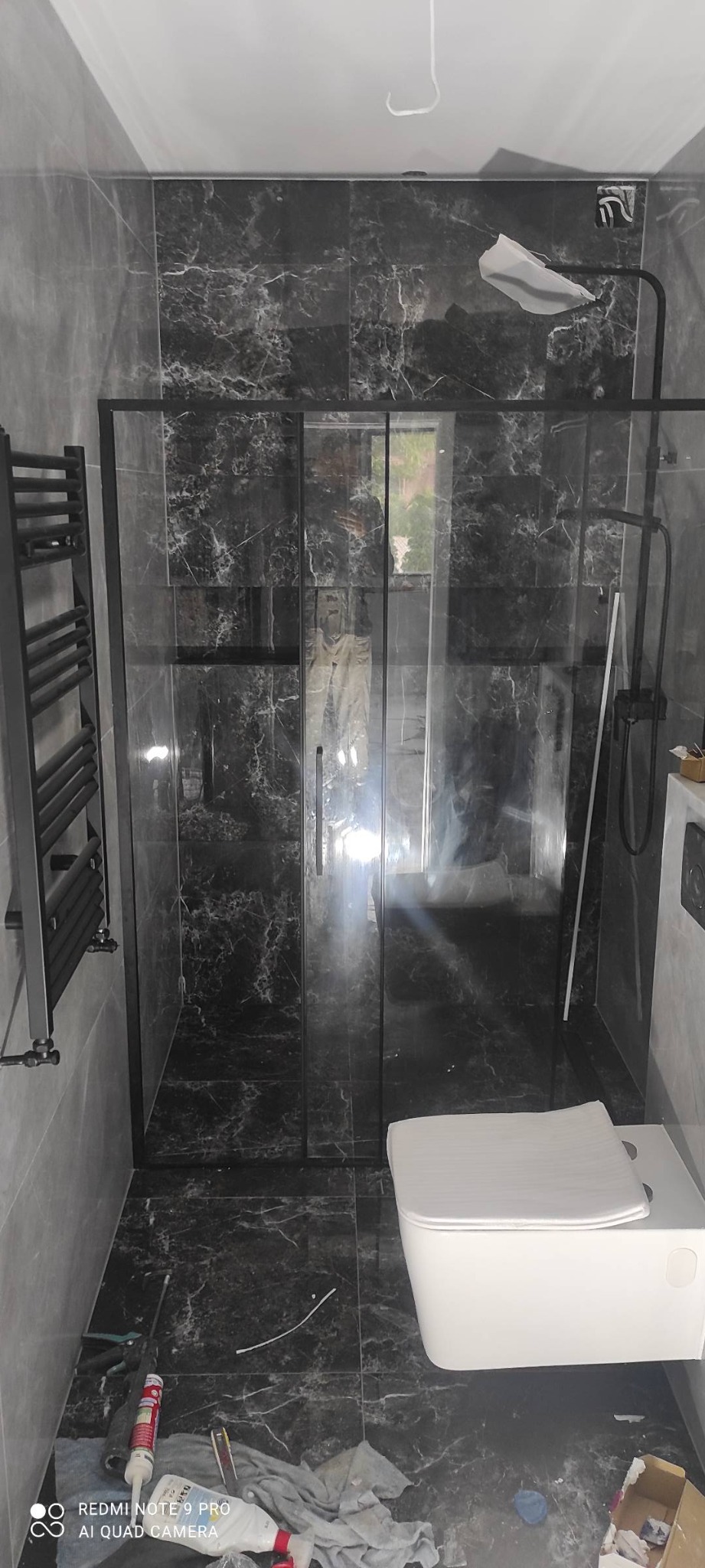 Nowoczesna łazienka z czarnymi płytkami imitującymi marmur, kabina prysznicowa z czarnymi ramami, wisząca toaleta i grzejnik drabinkowy, widoczne narzędzia i materiały budowlane na podłodze.