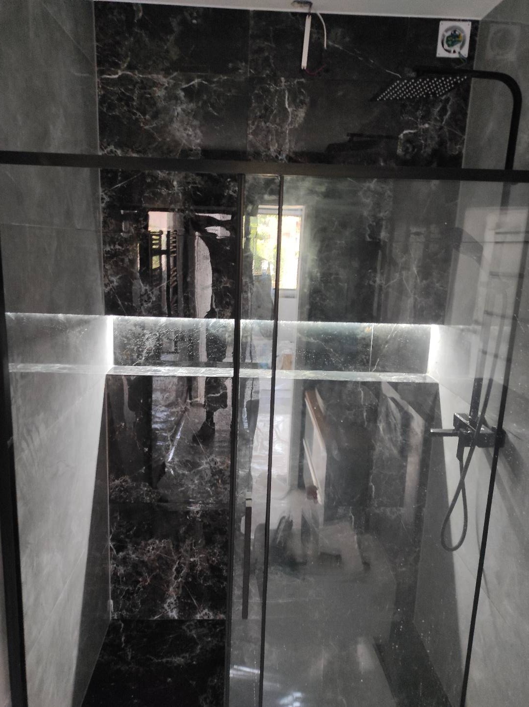 Nowoczesna kabina prysznicowa z czarnymi marmurkowymi płytkami i podświetleniem LED, widok przez szklane drzwi.