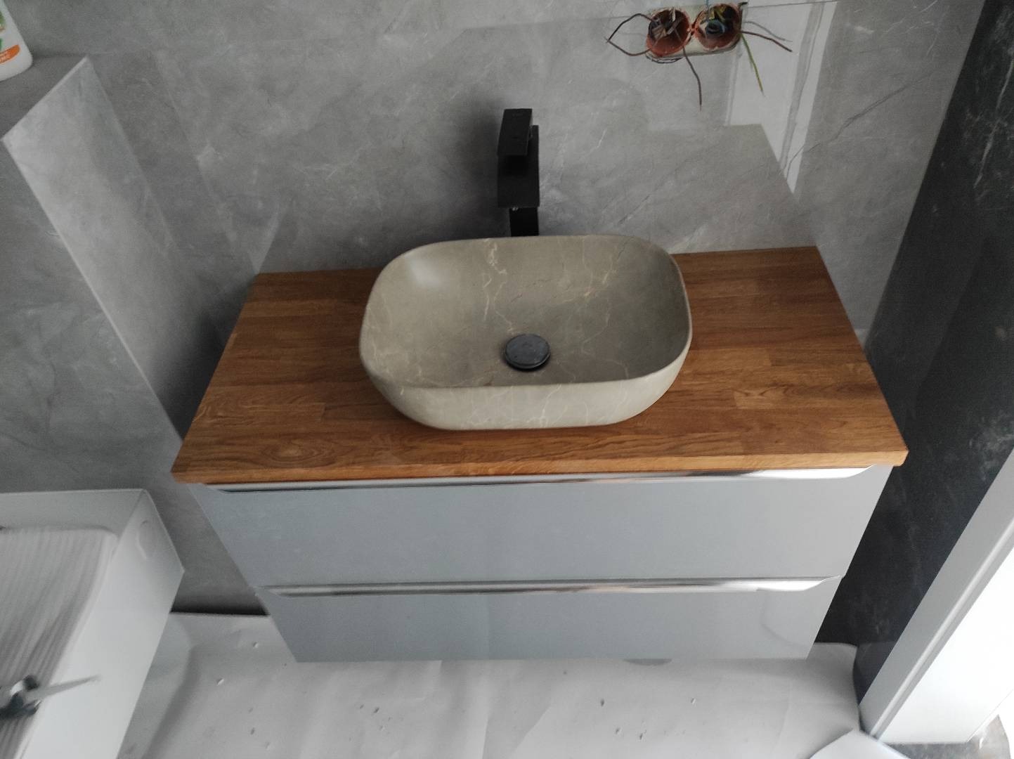 Beżowa umywalka nablatowa z marmurkowym wzorem, zamontowana na drewnianym blacie szafki łazienkowej z szufladami, na tle szarej ściany z widocznymi przewodami elektrycznymi.