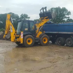 Żółto-czarna koparko-ładowarka JCB ładuje urobek do niebieskiej ciężarówki na rozmokłym placu budowy, w tle widoczny hałda materiału i zieleń drzew