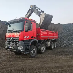 Czerwona ciężarówka Mercedes-Benz z otwartą skrzynią ładunkową, na którą koparka Volvo nabiera żwir z hałdy, widoczny numer rejestracyjny ZS 068TU, teren błotnisty.