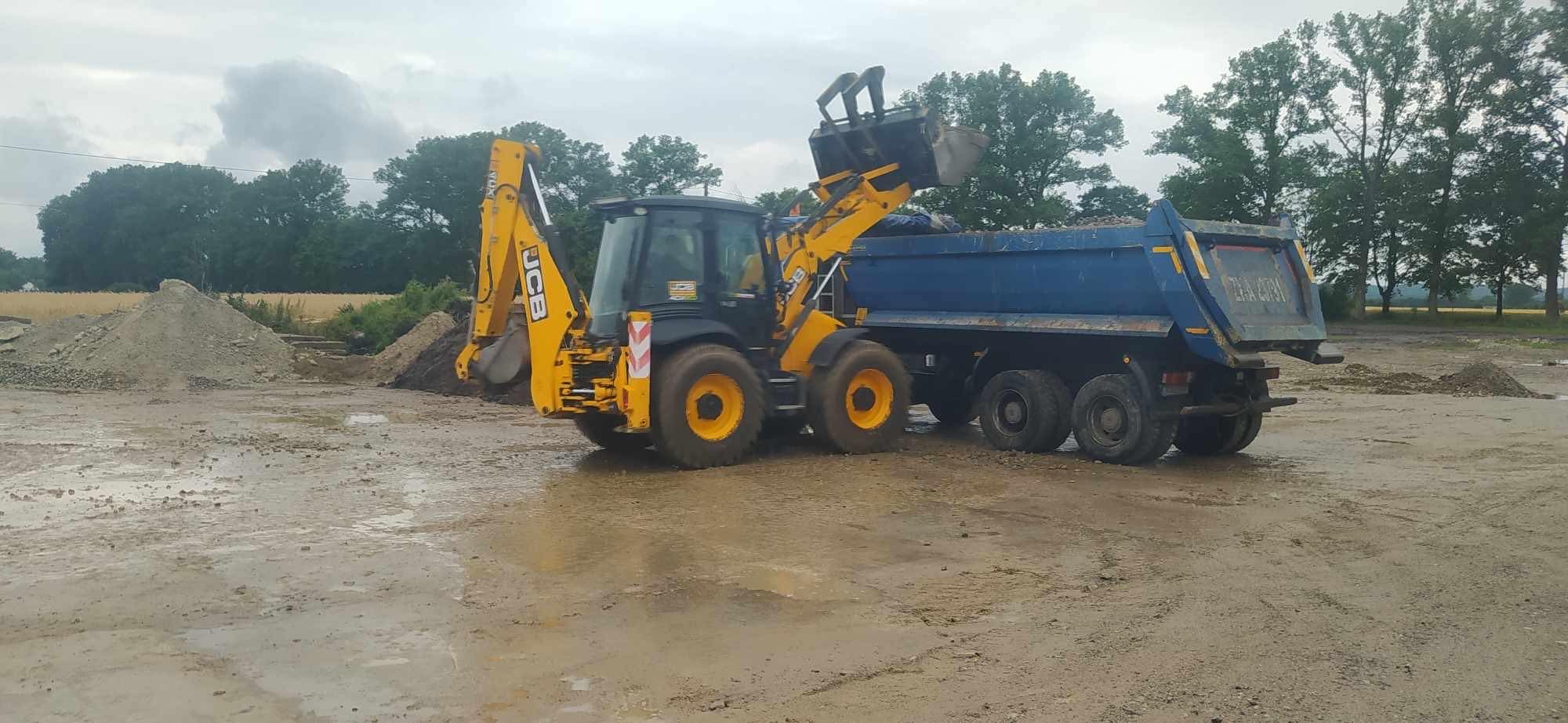 Żółto-czarna koparko-ładowarka JCB ładuje urobek do niebieskiej ciężarówki na rozmokłym placu budowy, w tle widoczny hałda materiału i zieleń drzew