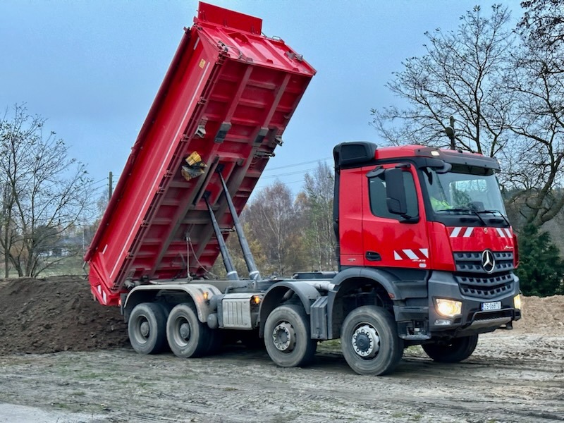 Czerwona wywrotka Mercedes z podniesioną skrzynią ładunkową wysypuje ziemię na placu budowy, widoczne drzewa w tle.