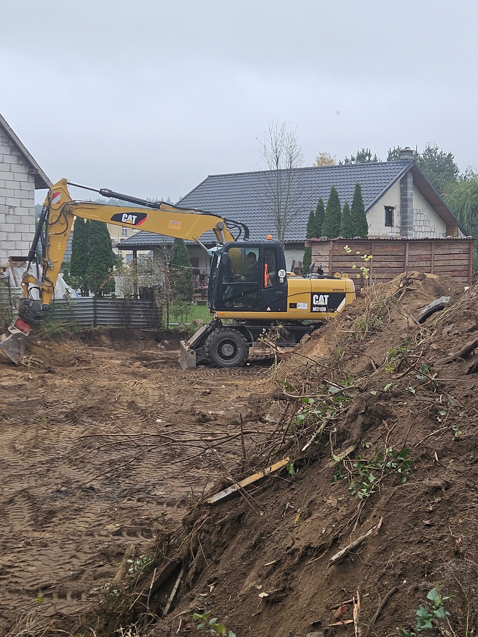 Żółta koparka CAT M315D w trakcie prac ziemnych na działce, widoczne ślady opon i wykop, w tle domy jednorodzinne i pochmurne niebo.