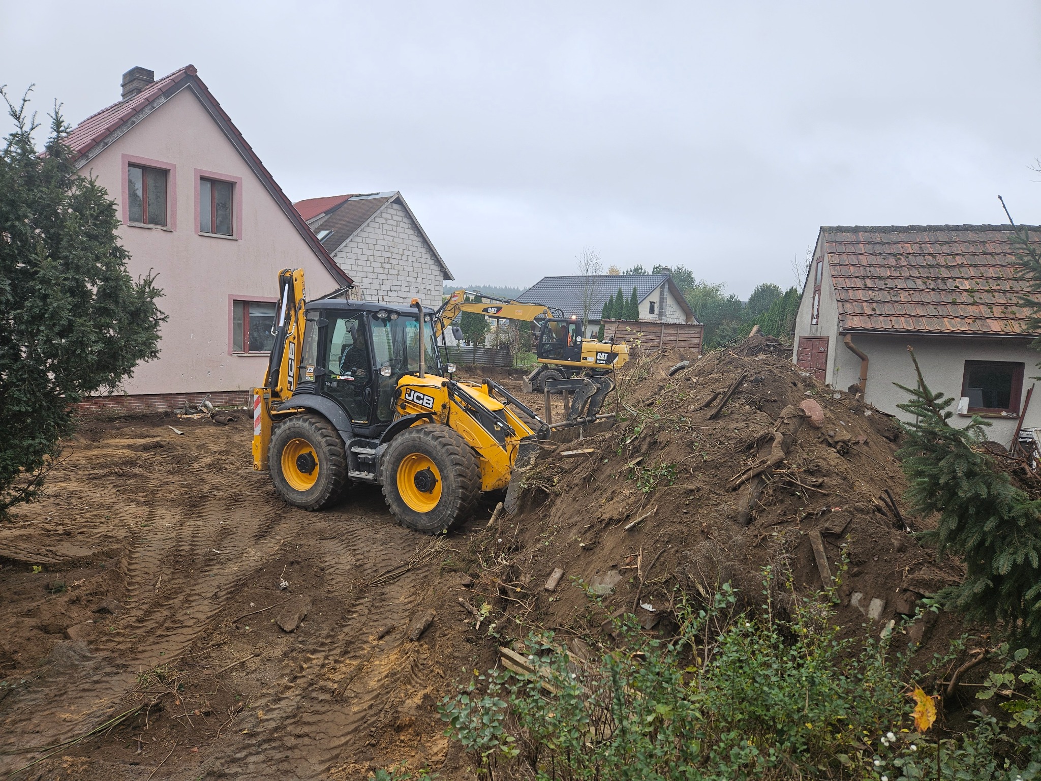 Dwa żółte ciągniki JCB i CAT pracują na placu budowy, jeden z nich nabiera ziemię z hałdy, w tle domy jednorodzinne w pochmurny dzień.