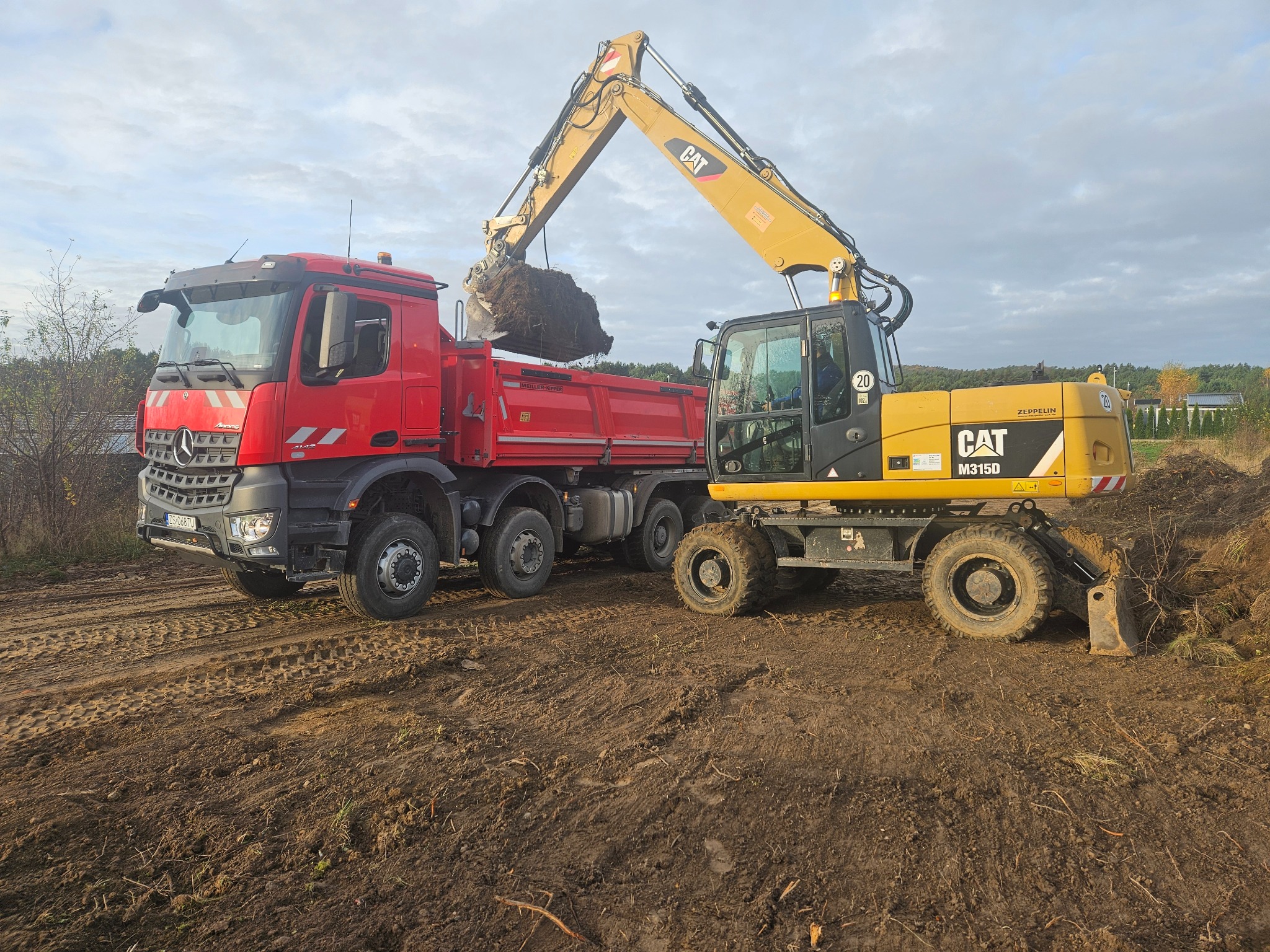 Żółta koparka CAT M315D nabiera ziemię i wrzuca ją do czerwonej ciężarówki Mercedes-Benz Actros na placu budowy, widoczne ślady opon na błotnistym podłożu.