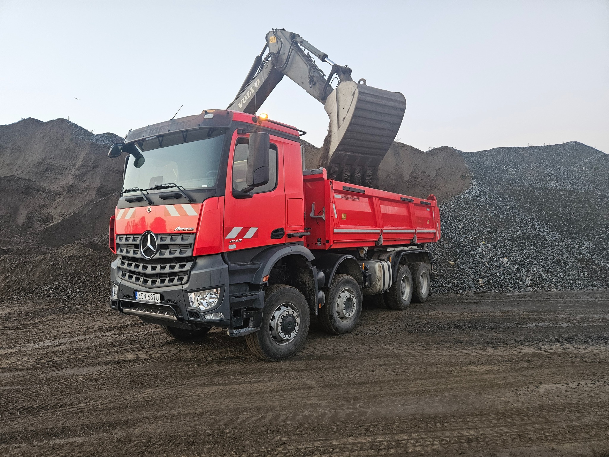Czerwona ciężarówka Mercedes-Benz z otwartą skrzynią ładunkową, na którą koparka Volvo nabiera żwir z hałdy, widoczny numer rejestracyjny ZS 068TU, teren błotnisty.