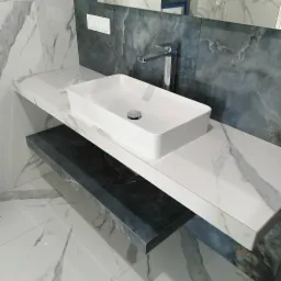 Nowoczesna, prostokątna umywalka nablatowa z chromowaną baterią na blacie z białego marmuru. Tło z płytek imitujących ciemny marmur. Eleganckie wykończenie łazienki.