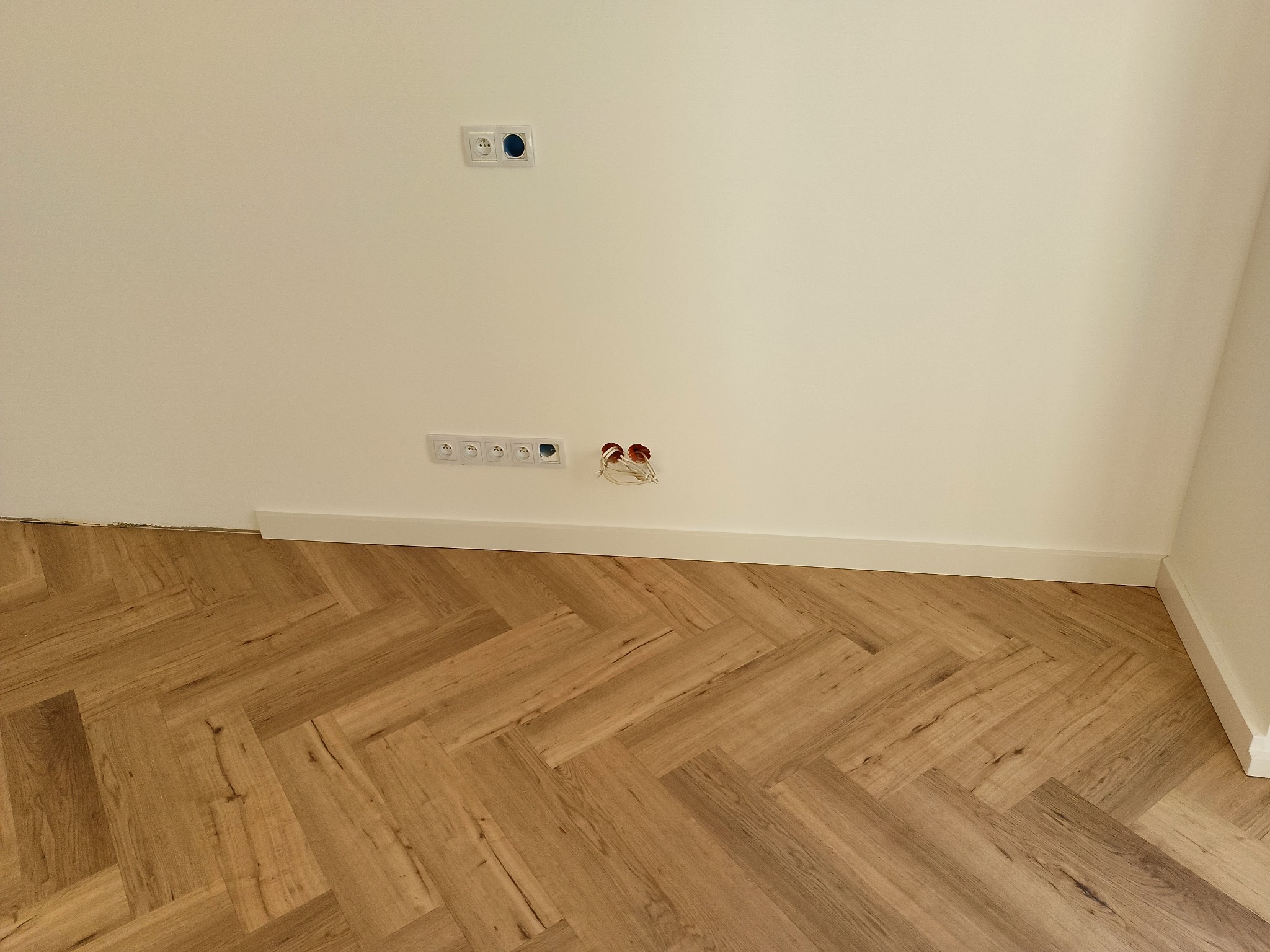 Jasna podłoga ułożona w jodełkę, białe ściany z listwami przypodłogowymi i gniazdkami elektrycznymi. Częściowo widoczne kable elektryczne.