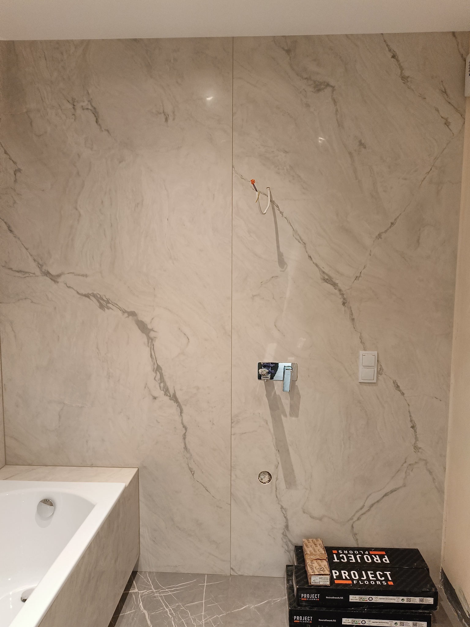 Ściana łazienki wyłożona wielkoformatowymi płytami imitującymi marmur, z widoczną baterią i włącznikiem światła. Na podłodze paczki paneli podłogowych.