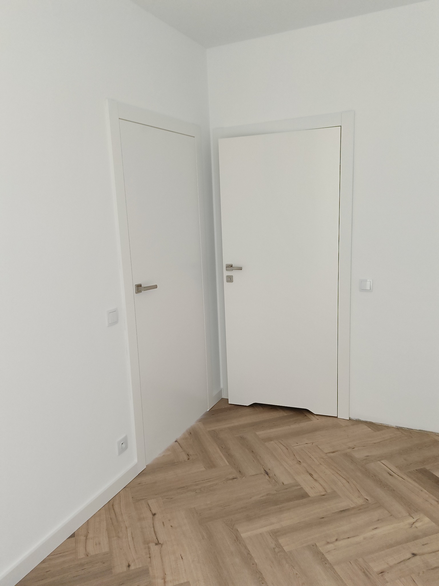 Dwie białe, minimalistyczne drzwi wewnętrzne z srebrnymi klamkami w nowocześnie wykończonym wnętrzu z parkietem ułożonym we wzór jodełki.