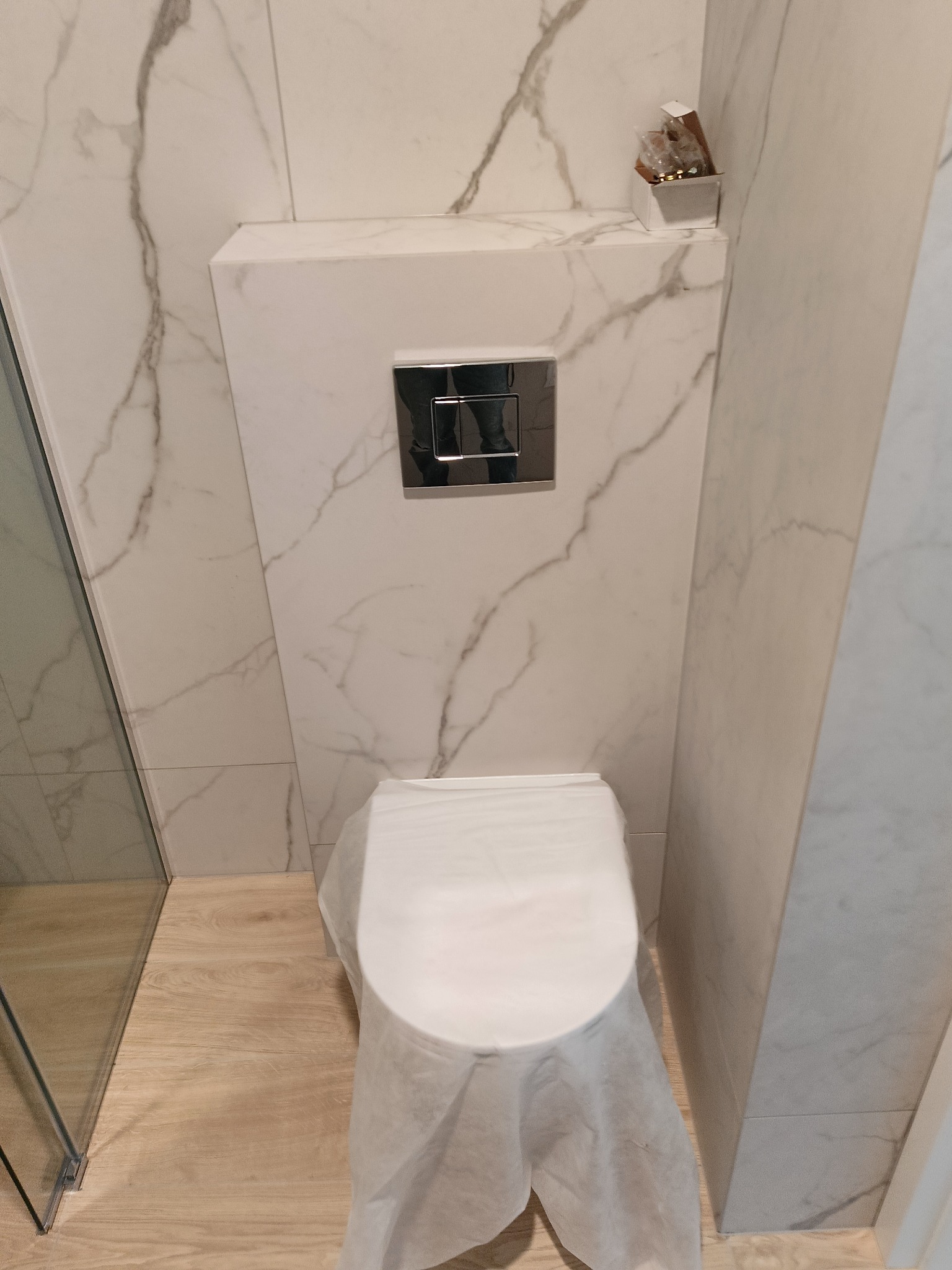 Nowoczesna toaleta podtynkowa z marmurową obudową i chromowanym przyciskiem, osłonięta folią ochronną, po remoncie, w jasnej łazience z drewnopodobną podłogą.