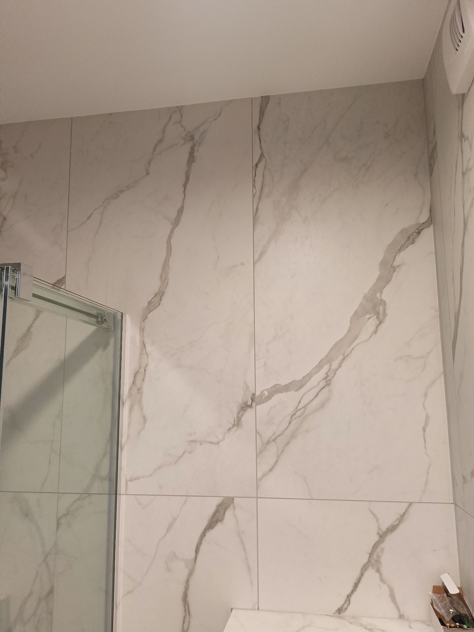 Wykończona ściana kabiny prysznicowej z płytek imitujących marmur z widocznym wentylatorem i fragmentem kabiny. Nowoczesny design łazienki po remoncie.