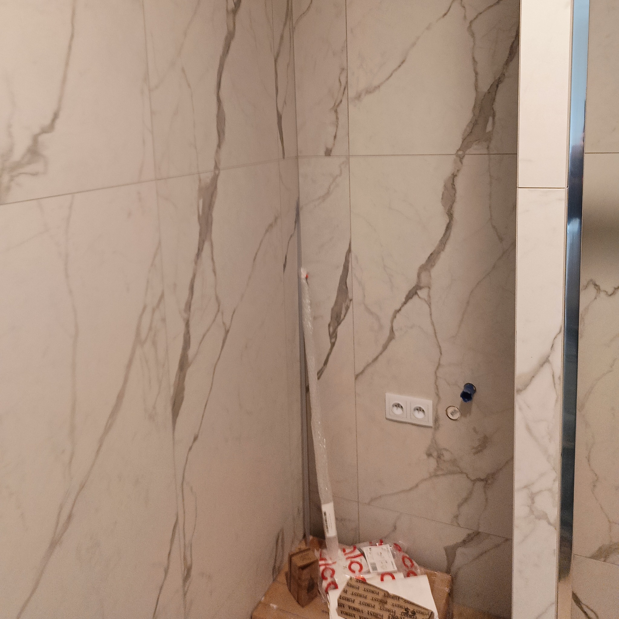 Wykończona ściana łazienki z marmurowymi płytkami, widoczna instalacja elektryczna i fragment pudełek z materiałami budowlanymi w rogu.