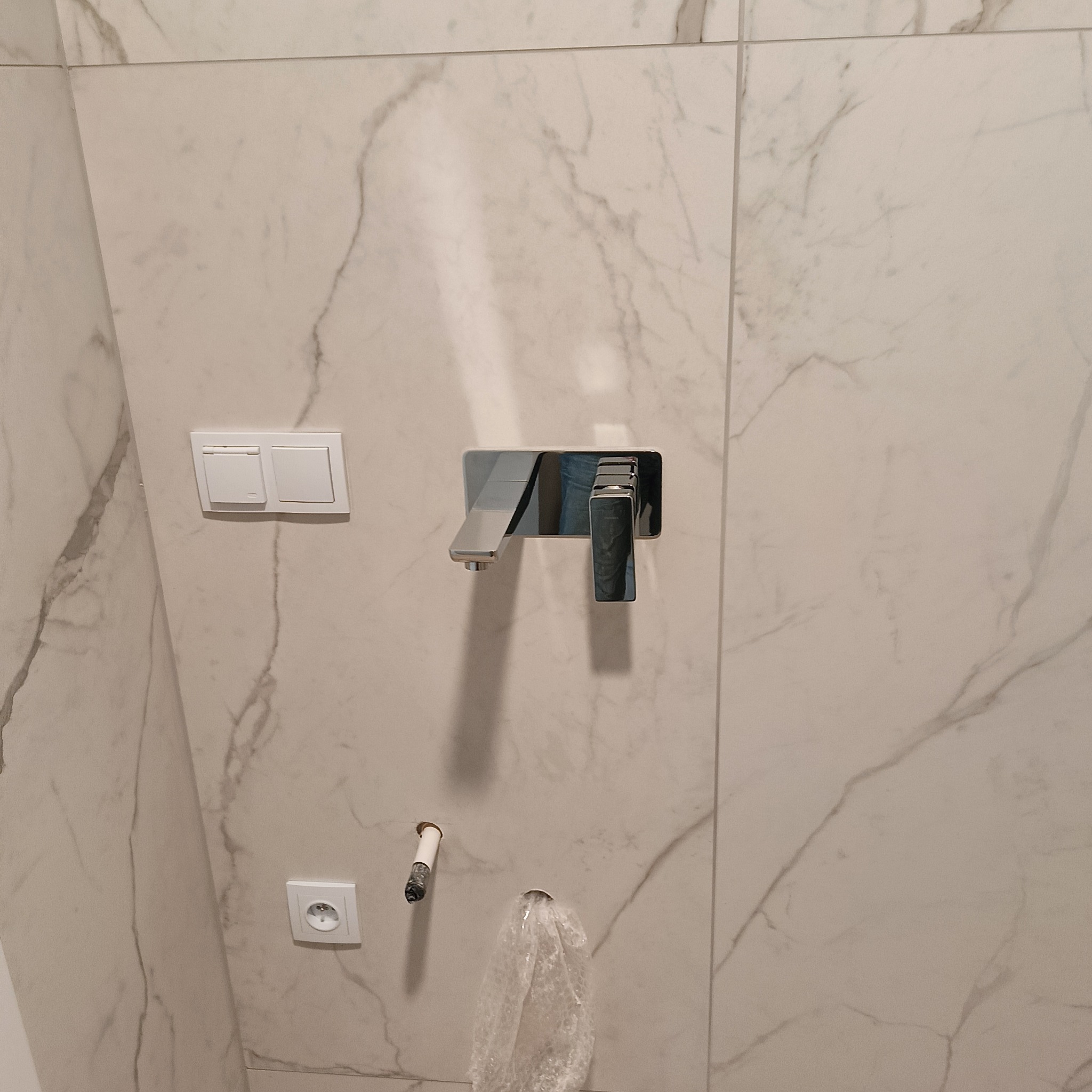 Biała ściana łazienki wyłożona płytkami imitującymi marmur, z zamontowaną nowoczesną, chromowaną baterią umywalkową i osprzętem elektrycznym. Widoczne przyłącza wodne.