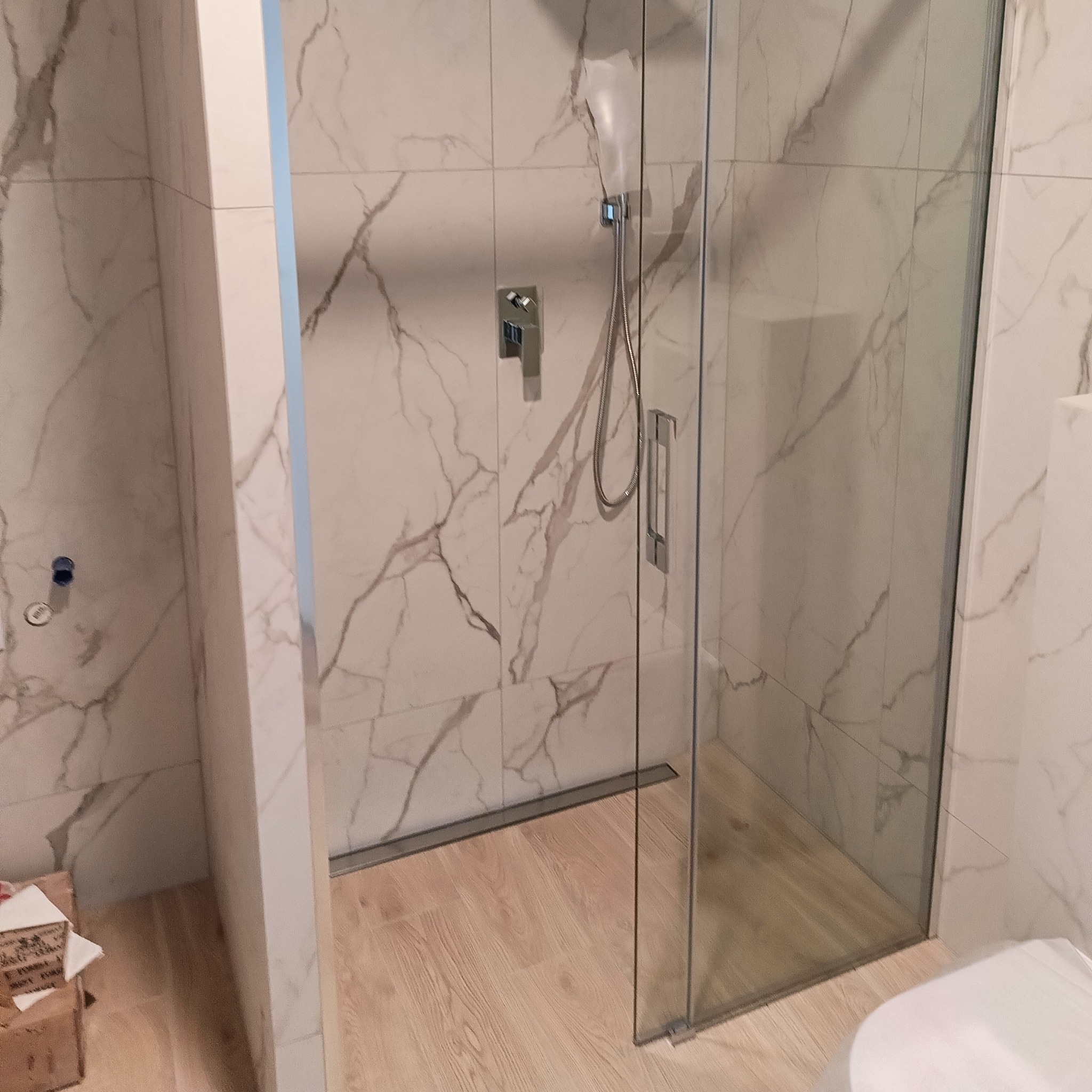 Wykończona kabina prysznicowa z deszczownicą i armaturą, ściany wyłożone płytkami imitującymi marmur, podłoga drewnopodobna, widoczny fragment toalety.