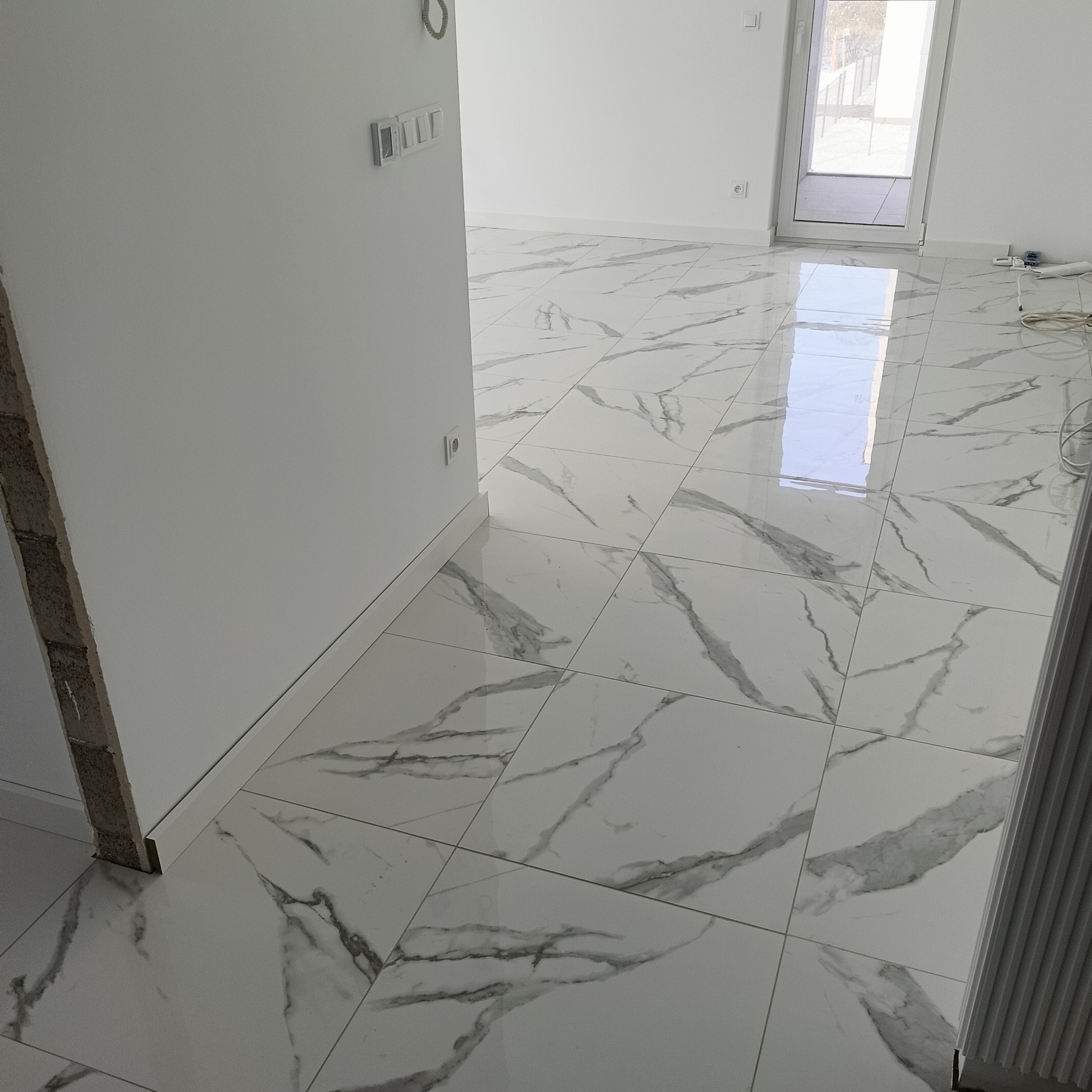 Białe płytki podłogowe z szarym wzorem marmuru w nowym, jasnym wnętrzu. Widoczne gniazdka elektryczne i fragment ściany w stanie surowym.