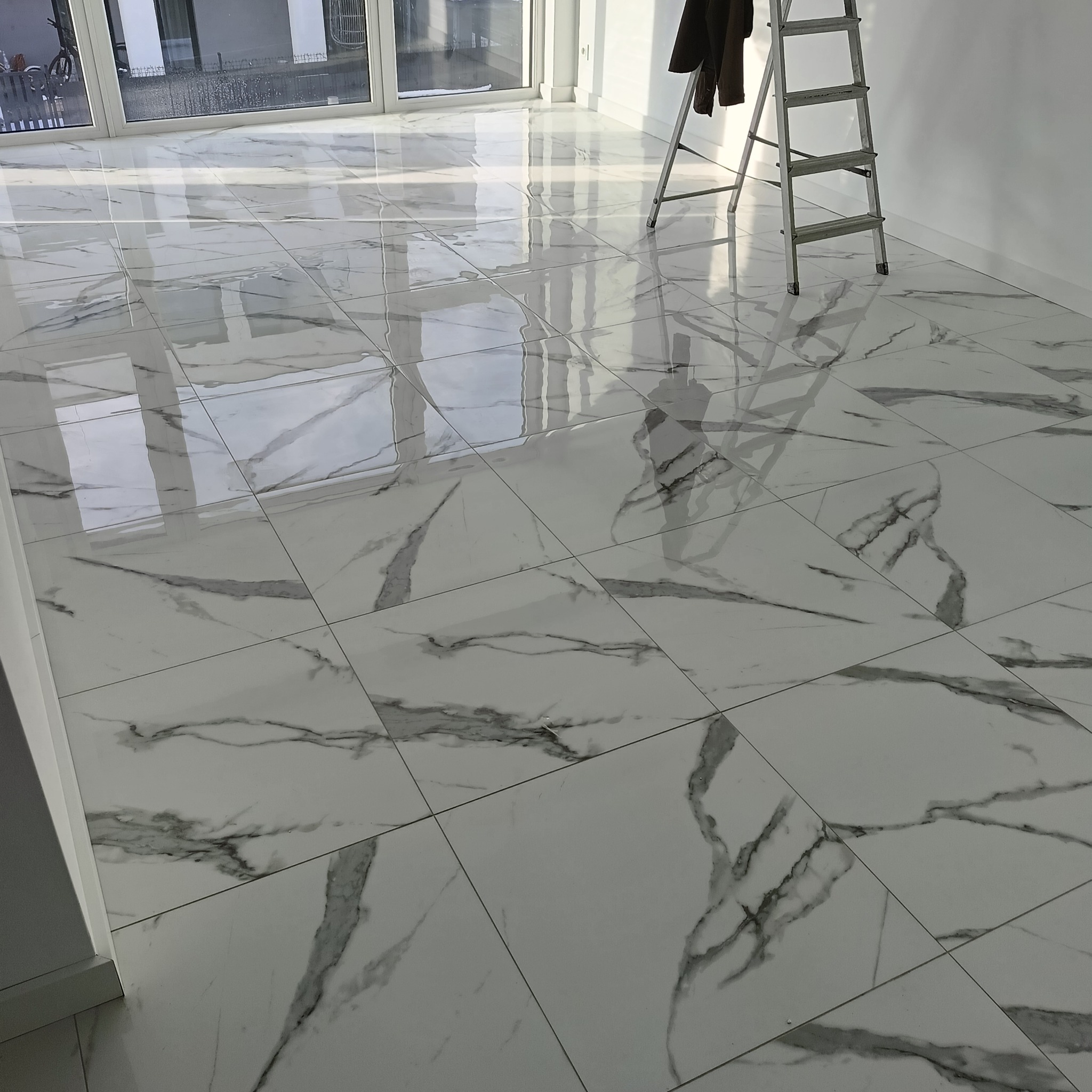 Połyskująca, biała podłoga z marmurowym wzorem, świeżo po ułożeniu. W tle okno balkonowe i drabina malarska. Widok na jasne, przestronne wnętrze.