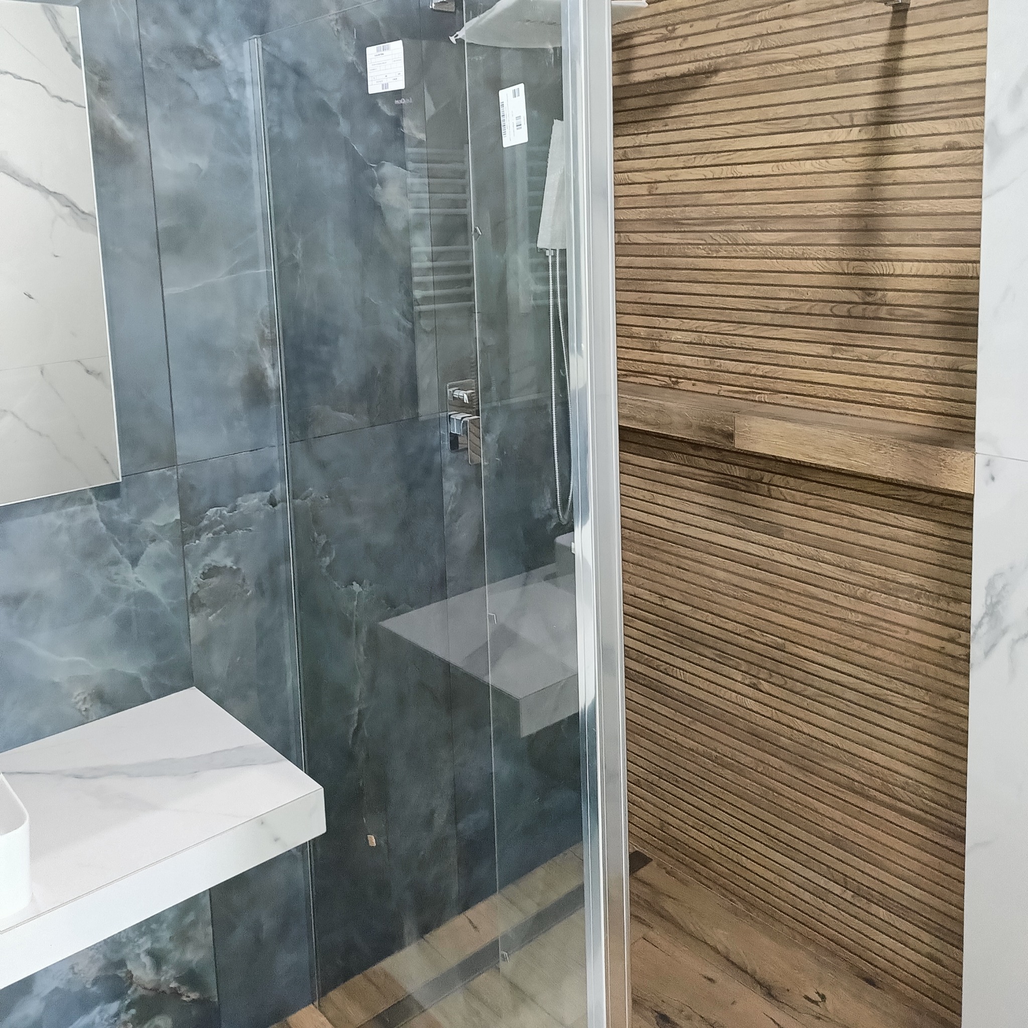 Nowoczesna kabina prysznicowa z szarymi płytkami imitującymi marmur i drewnianą ścianą. Umywalka z białym blatem. Minimalistyczny design, eleganckie wykończenie.