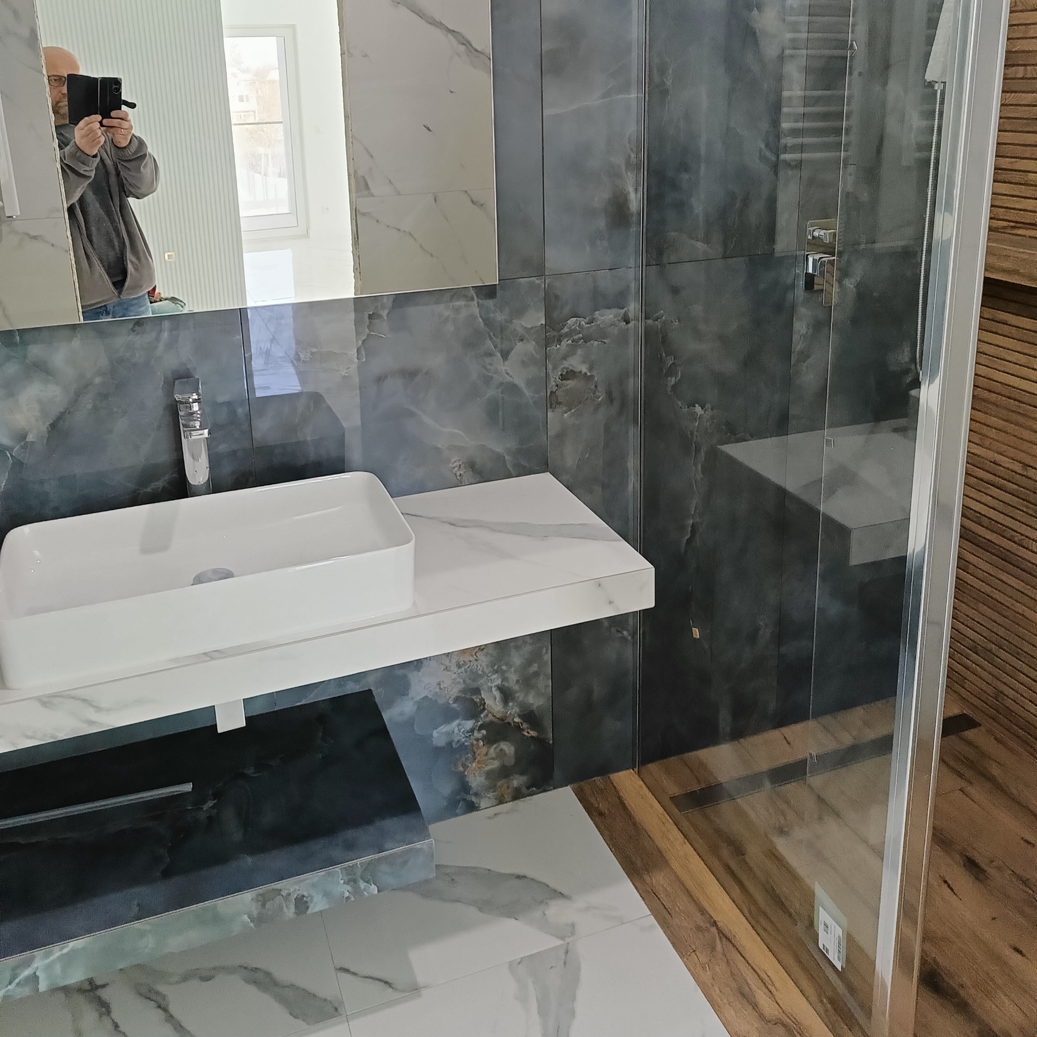 Nowoczesna łazienka z umywalką nablatową, baterią i lustrem. Szare płytki imitujące marmur, kabina prysznicowa ze szklaną ścianką. Widoczny fragment osoby w lustrze.