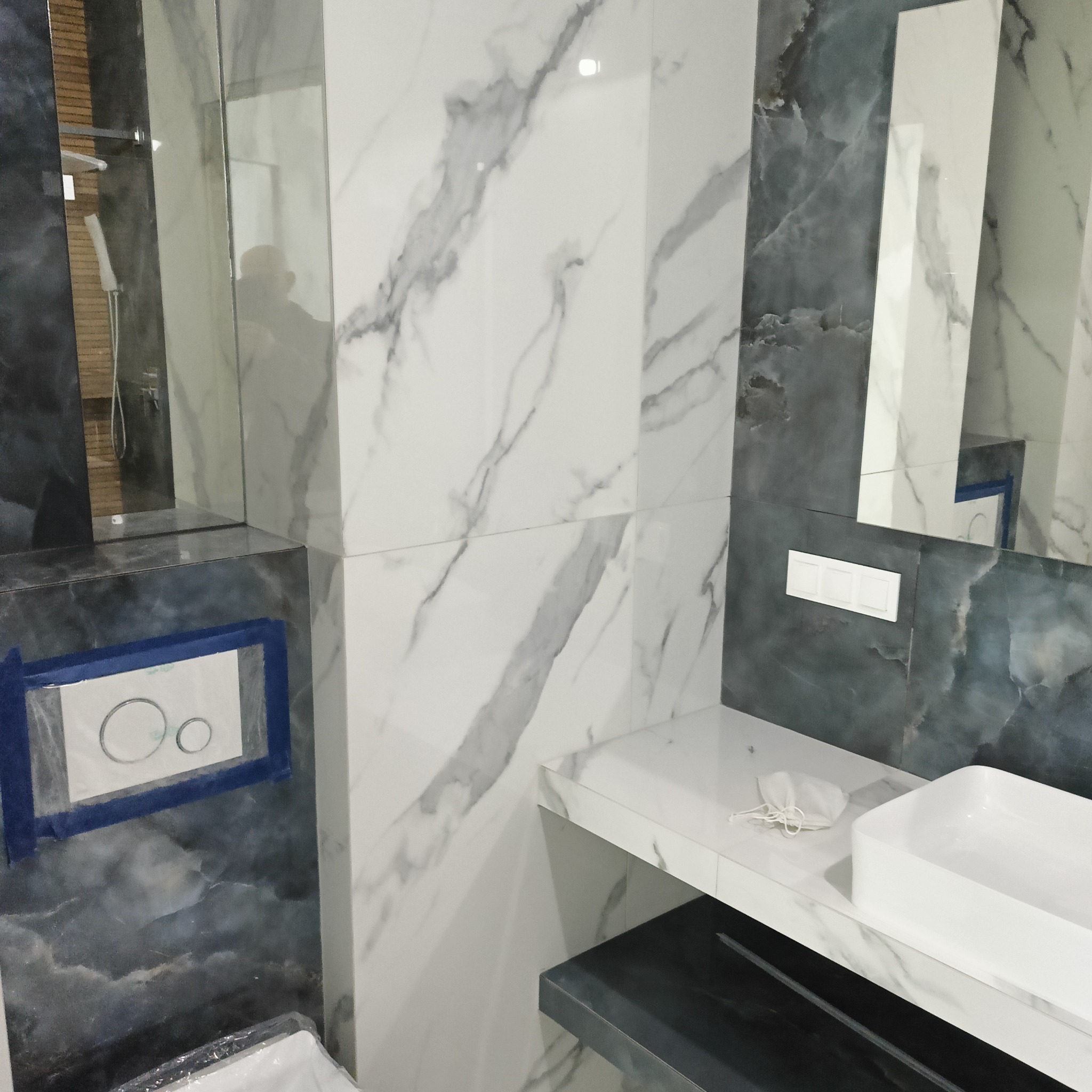 Nowoczesna łazienka z marmurowymi płytkami, białą umywalką i podtynkową spłuczką zabezpieczoną taśmą malarską. Widoczne odbicie sylwetki w lustrze.