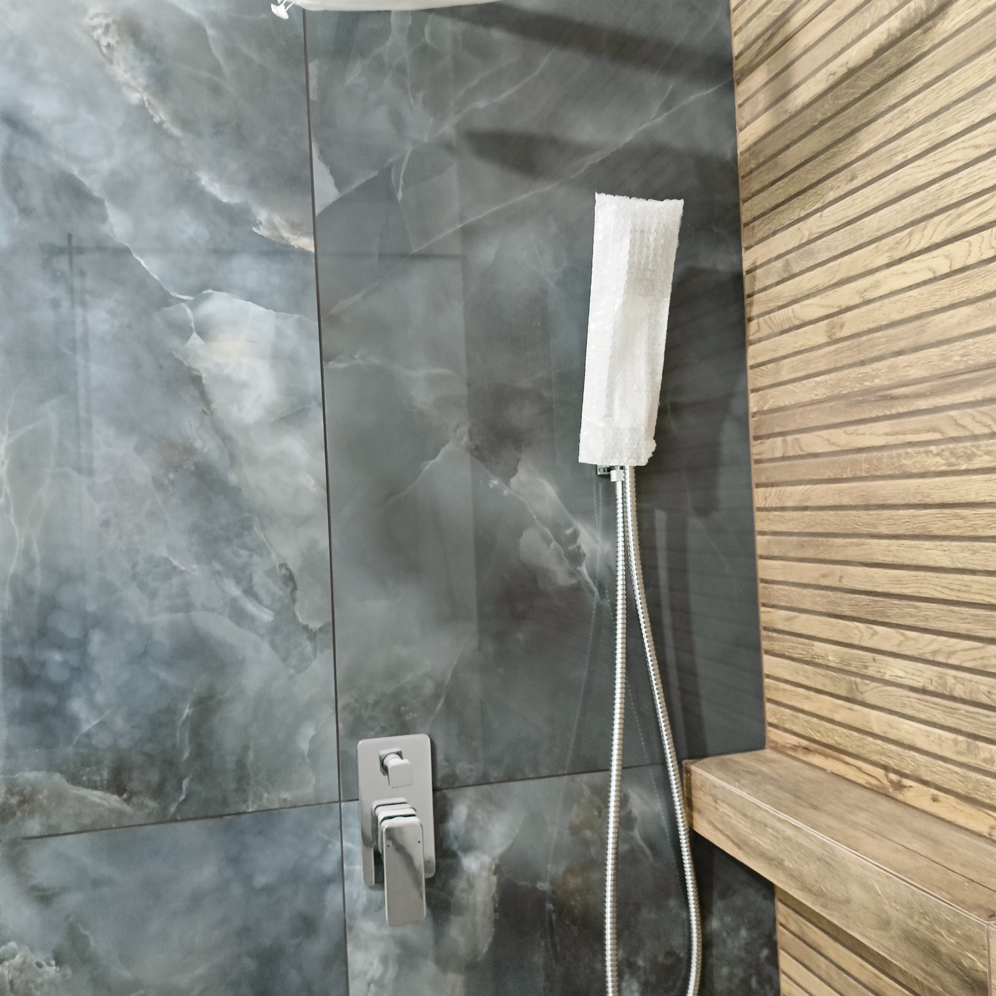 Nowoczesna łazienka z szarymi płytkami imitującymi marmur i drewnianą ścianą. Bateria prysznicowa z osłoną ochronną. Minimalistyczny design.