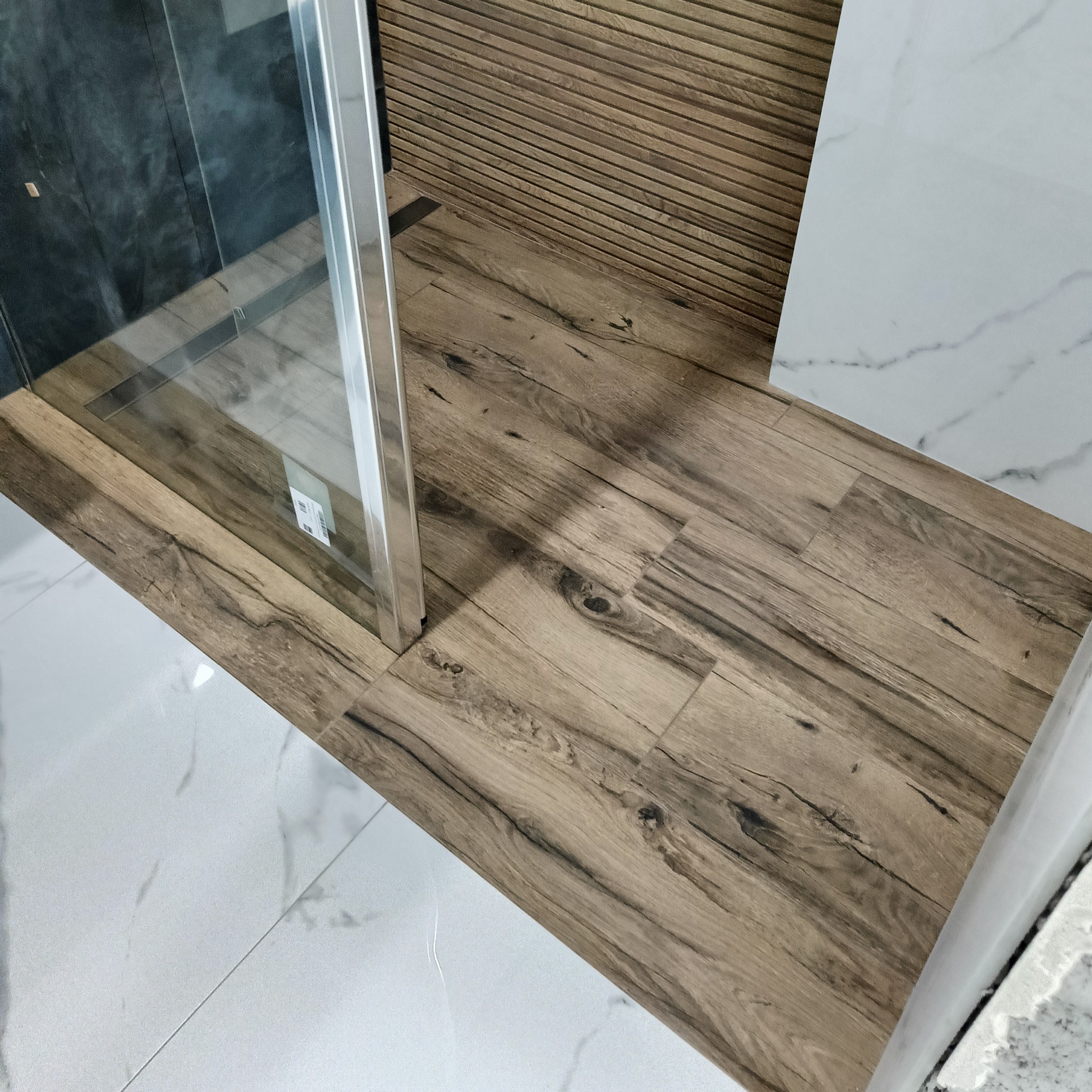 Nowoczesna łazienka po remoncie: drewnopodobne płytki pod prysznicem kontrastują z białym marmurem na podłodze i ścianie, widoczna kabina prysznicowa.