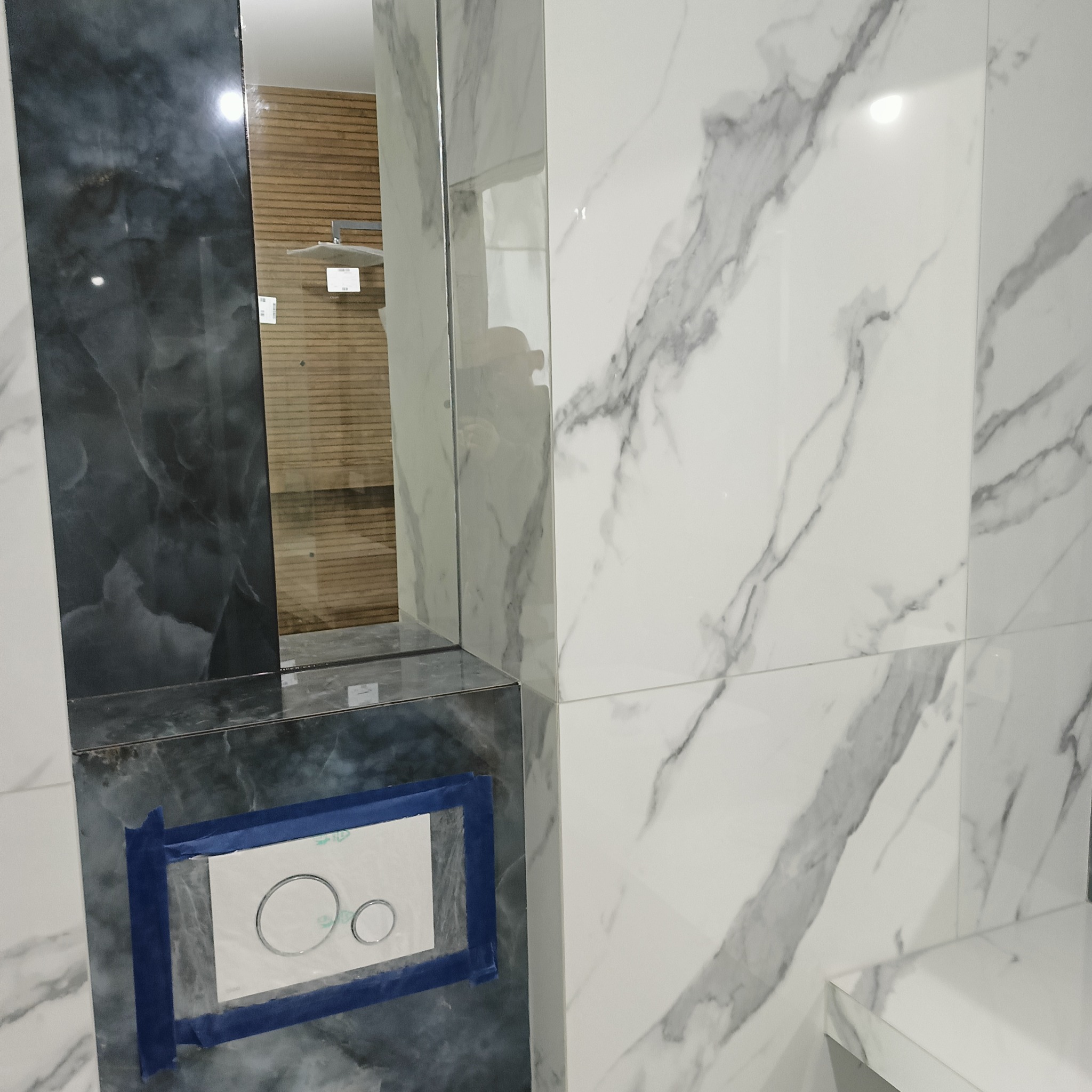 Wykończona łazienka z marmurowymi płytkami, widoczny fragment kabiny prysznicowej i podtynkowy stelaż WC z szablonem przycisku spłukującego przyklejonym taśmą.