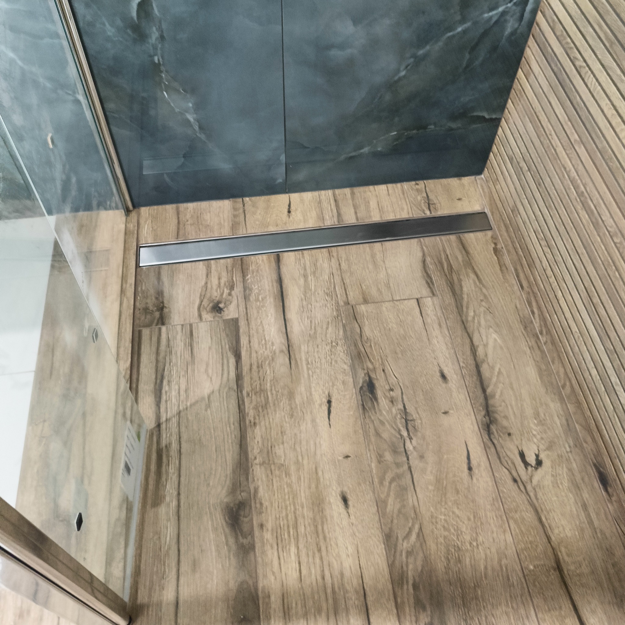 Nowoczesna kabina prysznicowa walk-in z odpływem liniowym. Drewnopodobne płytki podłogowe i szare płytki imitujące marmur na ścianach. Minimalistyczny design.