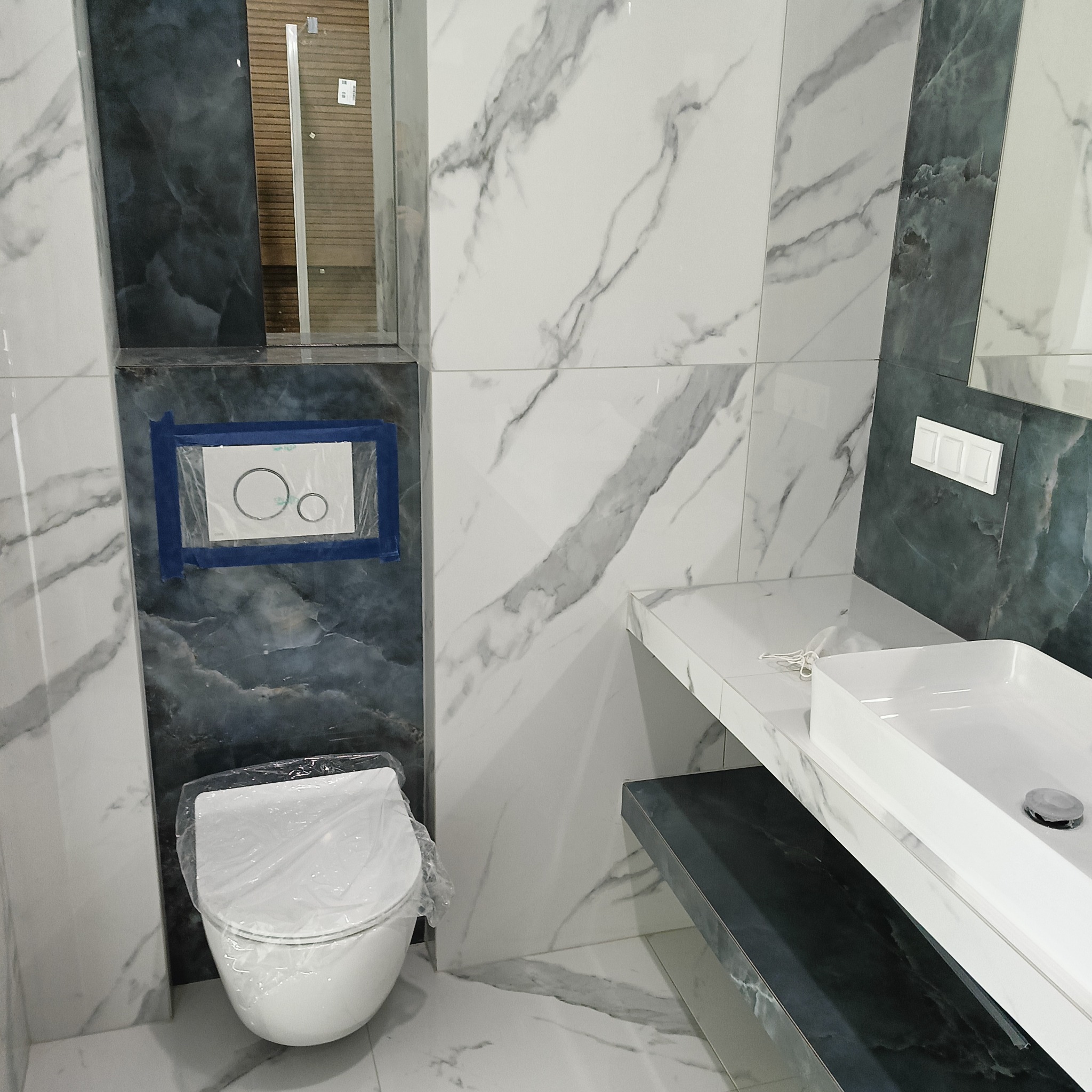 Nowoczesna łazienka w trakcie wykańczania: marmurowe płytki, podwieszana toaleta z zabezpieczeniem, umywalka nablatowa i elementy w odcieniach szarości.