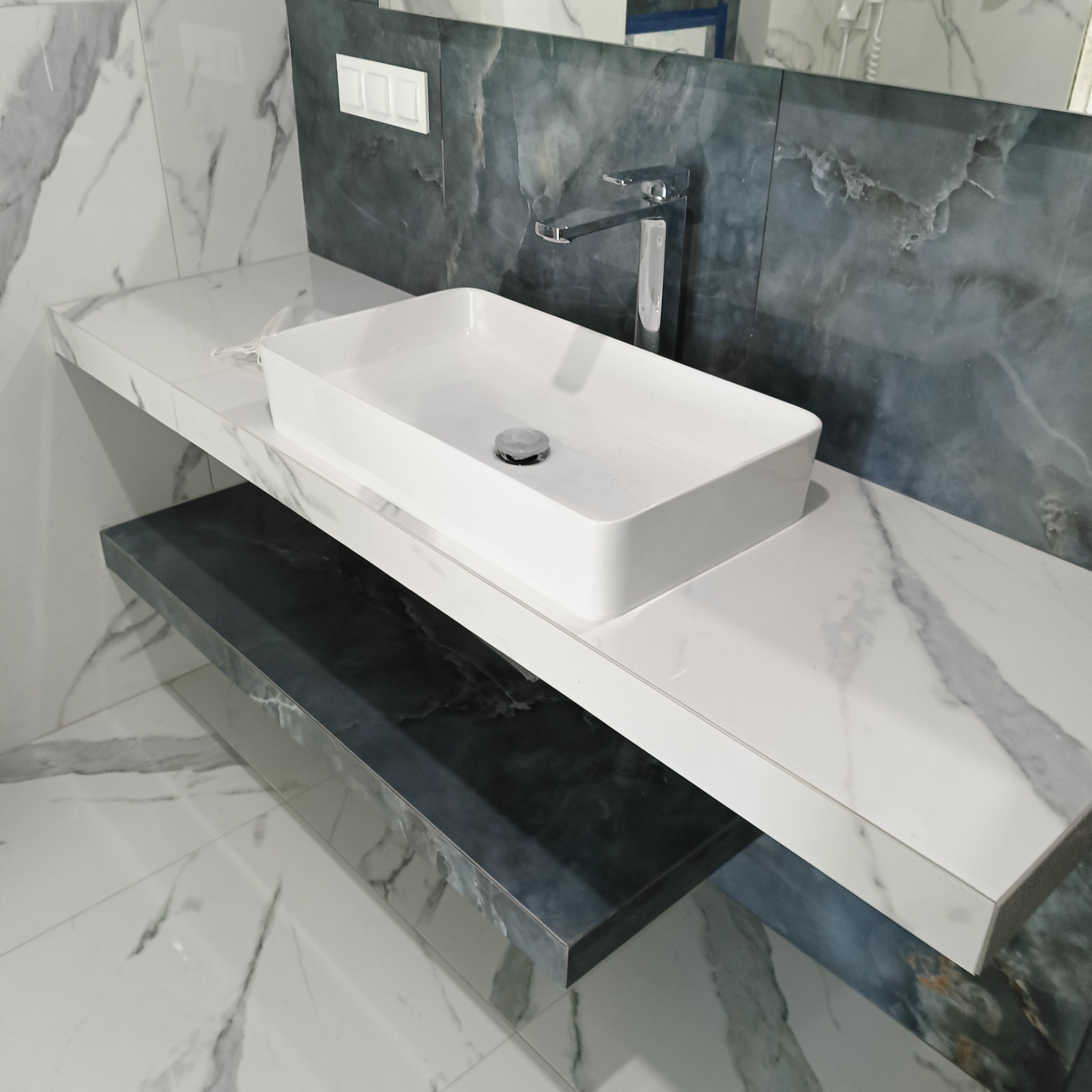 Nowoczesna, prostokątna umywalka nablatowa z chromowaną baterią na blacie z białego marmuru. Tło z płytek imitujących ciemny marmur. Eleganckie wykończenie łazienki.