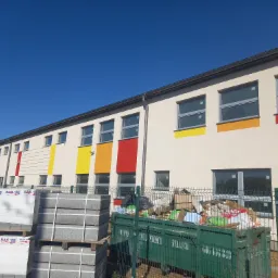 Nowo wybudowany budynek z beżową elewacją i kolorowymi pasami pod oknami, widoczne palety z kostką brukową i kontener ze śmieciami.