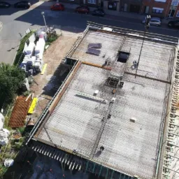 Widok z góry na plac budowy z przygotowaną konstrukcją zbrojeniową pod wylewkę fundamentów, widoczne deski szalunkowe, zbrojenie, wykop, w tle zabudowa miejska z cegły.