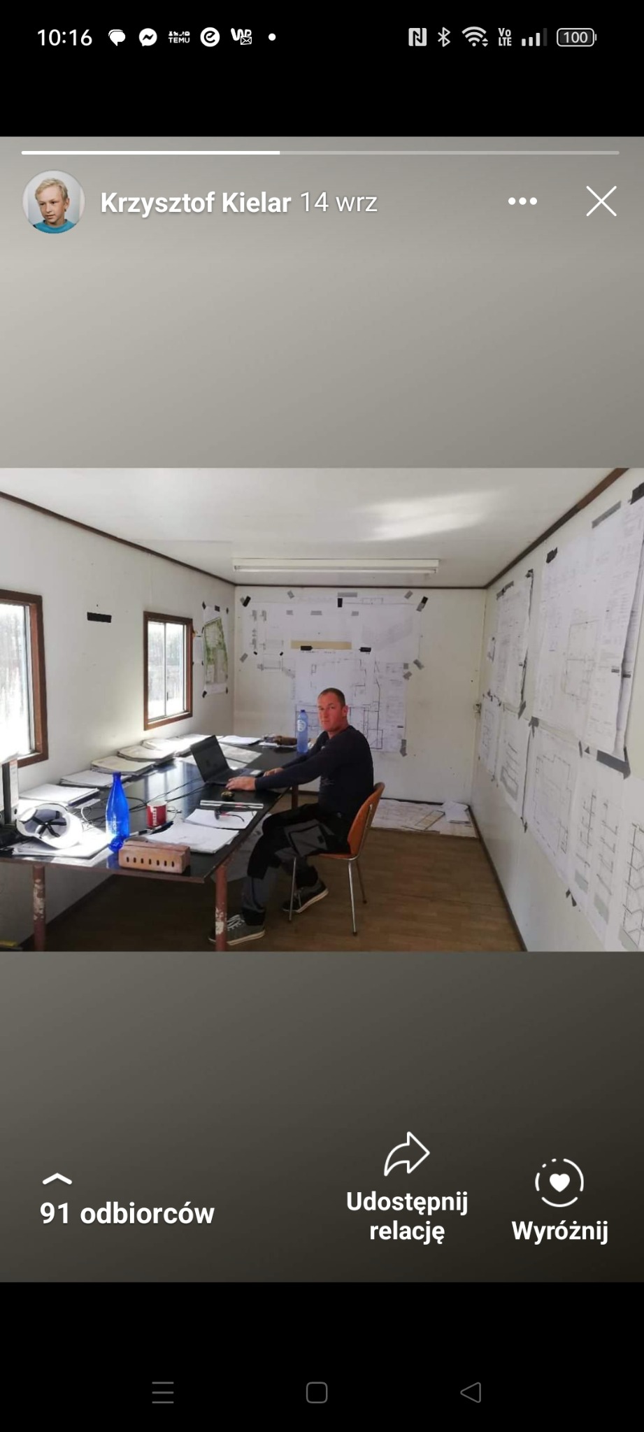 Mężczyzna siedzi przy długim stole w biurze kontenerowym, pracując na laptopie, otoczony planami budynków przyklejonymi do ścian.