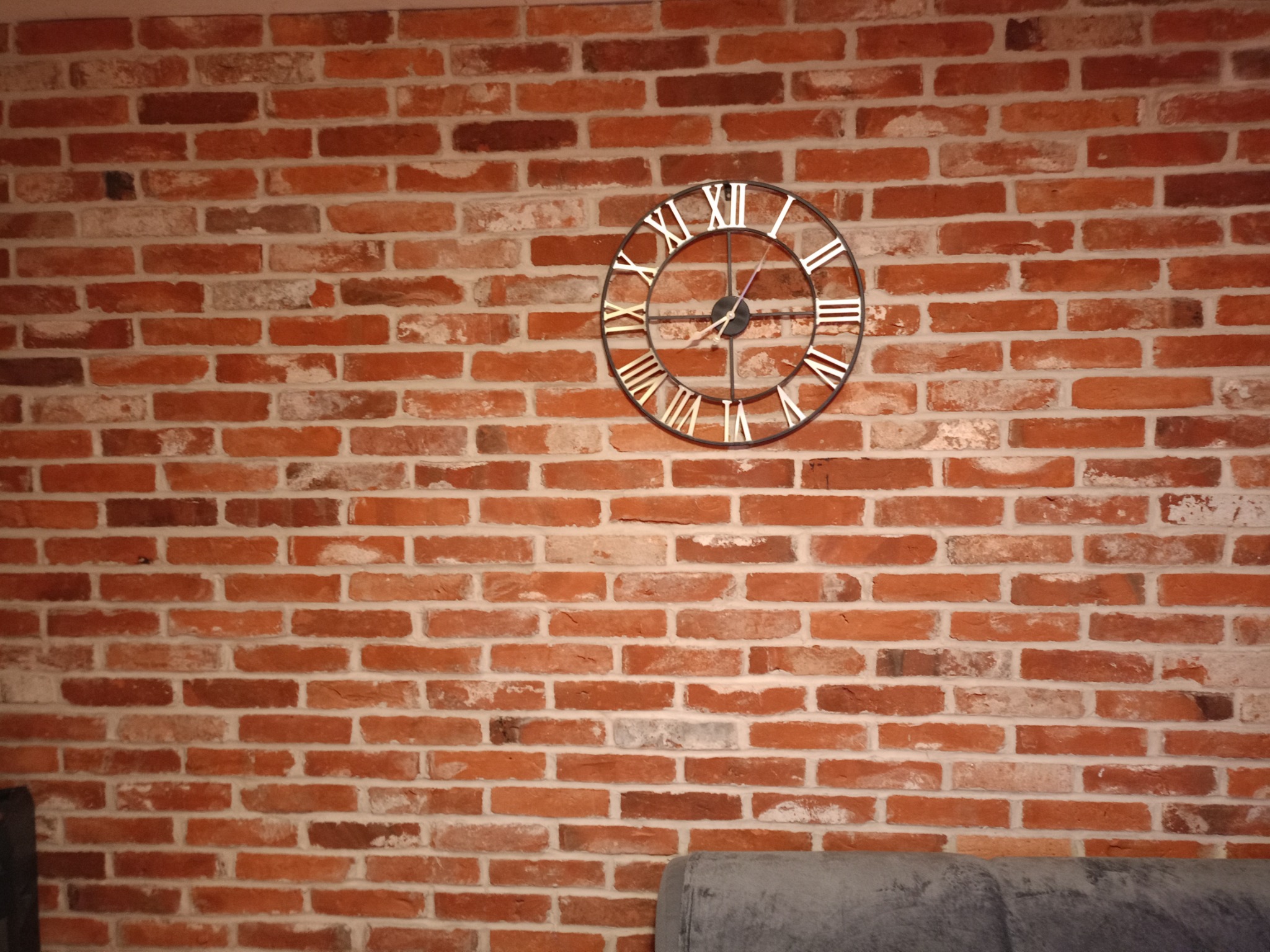 Dekoracyjna ściana z czerwonej cegły z metalowym zegarem w stylu retro z rzymskimi cyframi, fragment sofy w dolnym kadrze.