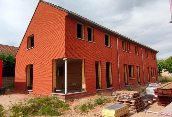 Czerwony, murowany budynek w stanie surowym otwartym, widoczne otwory okienne bez okien, fragment zadaszenia w trakcie budowy, palety z materiałami budowlanymi na pierwszym planie.