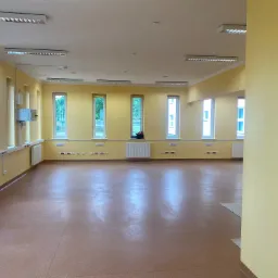 Pusta sala z żółtymi ścianami, brązową podłogą, białymi grzejnikami i oknami z widokiem na zieleń, oświetlona przez szereg lamp sufitowych.