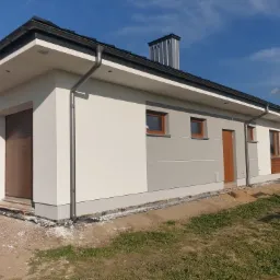 Nowoczesny parterowy dom z biało-szarą elewacją, brązowymi oknami i drzwiami, widoczne rynny, dach z kominem, teren wokół domu częściowo zagospodarowany.