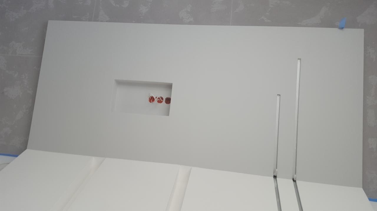 Biała ściana z wyciętym otworem na gniazdka elektryczne i dwoma pionowymi wgłębieniami, widoczne przewody, fragment podłogi zabezpieczony taśmą malarską.