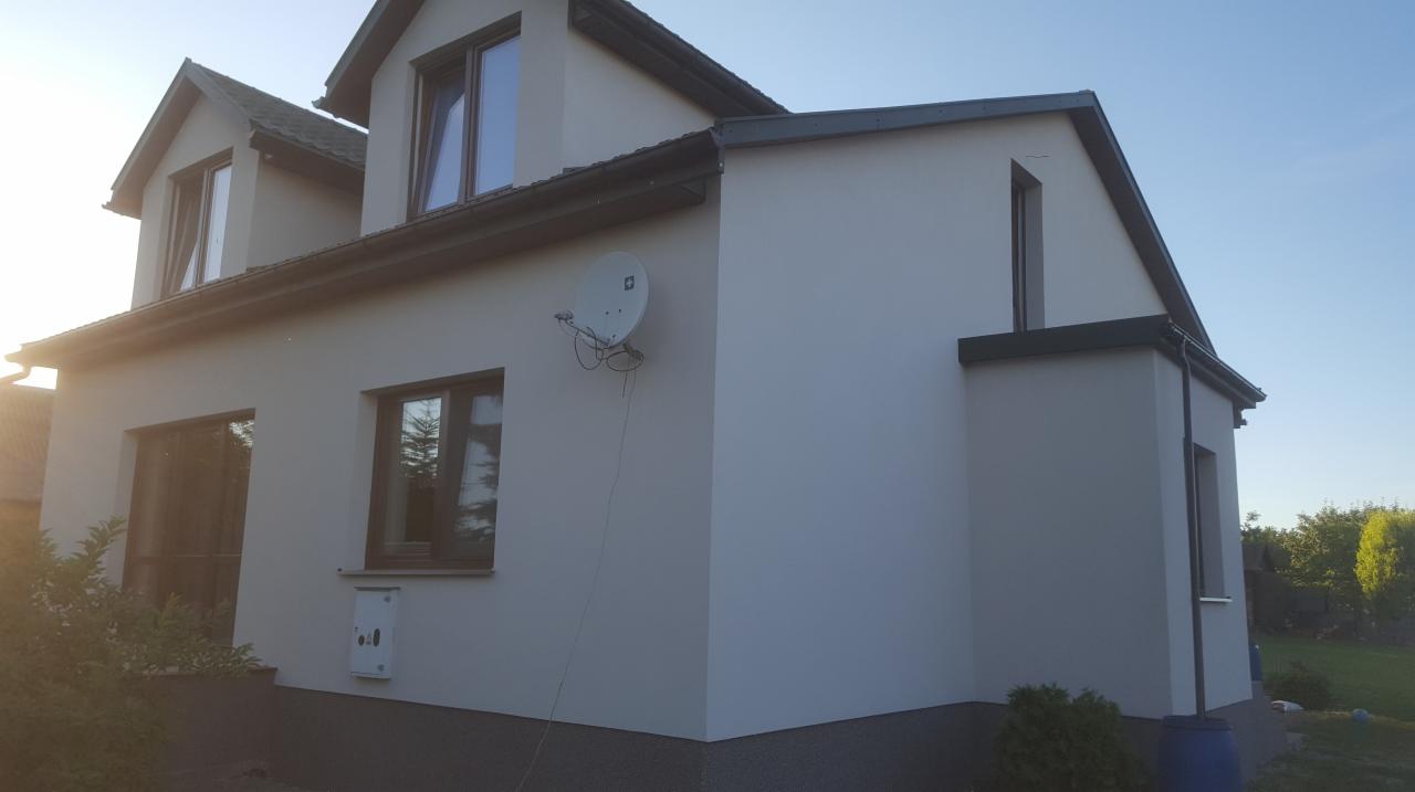 Jasnoszary dom jednorodzinny z ciemną stolarką okienną i anteną satelitarną na elewacji, widok z boku w słoneczny dzień.