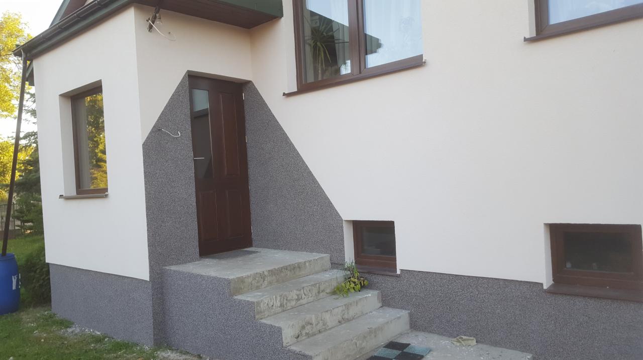 Wykończona elewacja domu z wejściem, schodami betonowymi i elementami dekoracyjnymi w kolorze szarym, okna z brązowymi ramami.