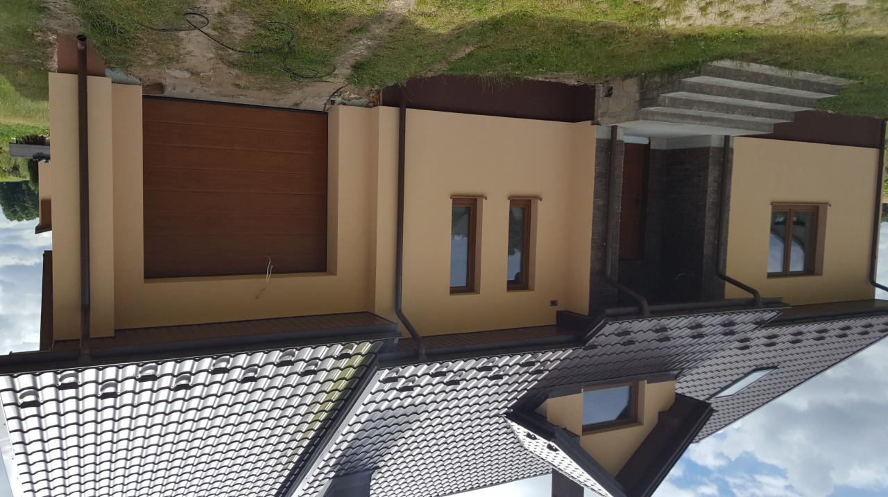 Jasna elewacja domu jednorodzinnego z brązowymi oknami i bramą garażową, widoczna z perspektywy dachu z dachówki ceramicznej.