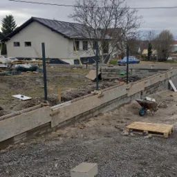 Fundamenty domu jednorodzinnego w trakcie budowy, widoczne szalunki, stalowe pręty zbrojeniowe, gruz, taczka, betoniarka i biały bus w tle.